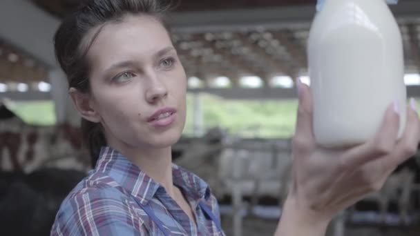 Retrato de una joven trabajadora en la granja de vacas comprobando la calidad de la leche en el primer plano de la botella. El agricultor positivo controla el proceso de trabajo. Industria agrícola, agricultura y ganadería — Vídeos de Stock