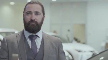 Fotoğraf makinesine bakarak lüks bir aracın anahtarını gösteren bir iş elbisesinden başarılı bir sakallı iş adamın portresi. Araba showroom. Reklam konsepti.