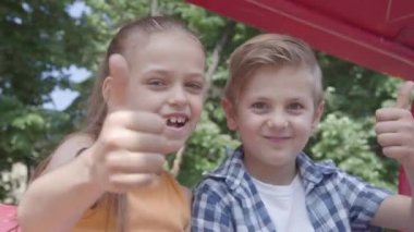 Sevimli erkek ve kız parkta yakın salıncak üzerinde oturan başparmak gösteren, kameraya bakarak. Birkaç mutlu çocuk. Aşık komik çocuklar.