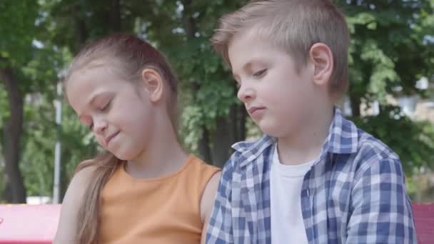 Portret van schattige blonde jongen zoenen Wang van een mooi meisje zittend op de schommel op de speelplaats. Het meisje is verlegen. Een paar gelukkige kinderen. Grappige kinderen verliefd — Stockvideo