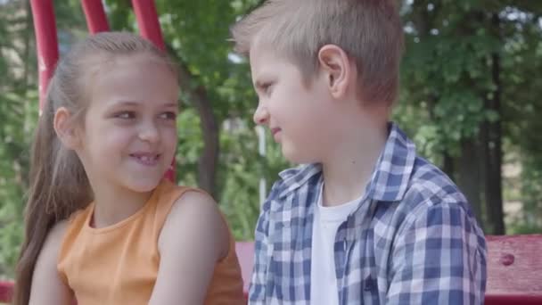 Retrato de chico rubio lindo besando mejilla de una chica bonita sentada en el columpio en el patio de recreo. La chica es tímida. Un par de niños felices. Niños divertidos en el amor — Vídeos de Stock