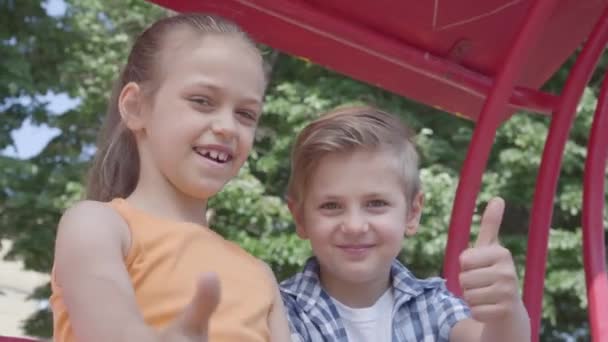 Schattige jongen en meisje tonen duimen omhoog zittend op de schommel close-up in het Park, kijkend naar de camera. Een paar gelukkige kinderen. Grappige kinderen verliefd. — Stockvideo