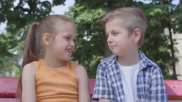 Bedårande pojke och söt flicka sitter på gungan närbild i parken, prata och dansa, ha kul. Ett par glada barn. Rolig bekymmerslös barn i kärlek utomhus. — Stockvideo