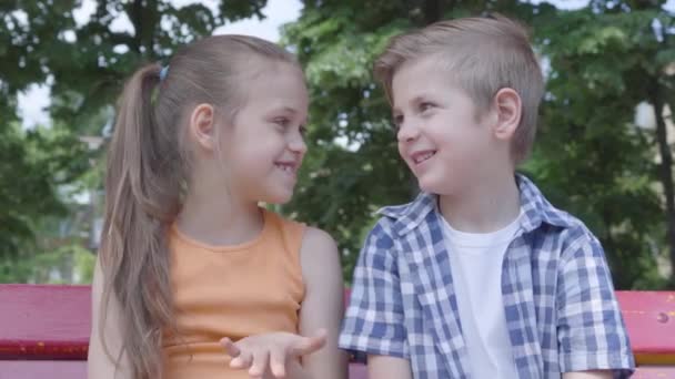 Portrait mignon garçon et fille assis sur la balançoire près du parc, dansant, s'amusant à regarder à la caméra. Deux enfants heureux. drôles insouciants enfants en amour à l'extérieur . — Video