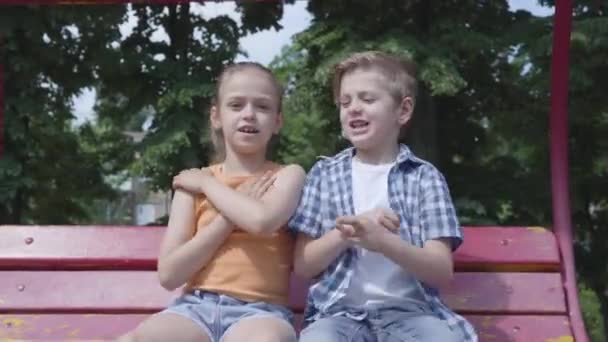 Urocza chłopiec i cute girl siedzi na huśtawce z bliska w parku, taniec, zabawy patrząc w aparacie na zewnątrz. Kilka szczęśliwych dzieci. Śmieszne beztrodarmowe dzieci w miłości — Wideo stockowe