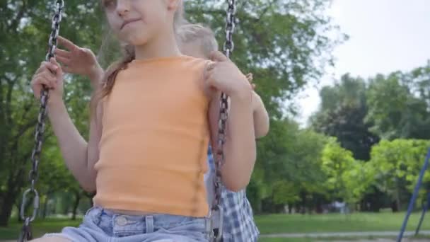 Lindo chico balanceándose en un columpio hermosa chica con el pelo largo, sonriendo. Un par de niños felices. Divertidos niños despreocupados en el amor . — Vídeo de stock