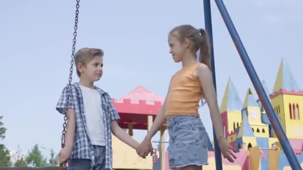 Little boy and beautiful girl with long hair holding hands and talking near the swing, smiling happily. A couple of happy children. Funny carefree kids in love. Boy courting girl — Stock Video