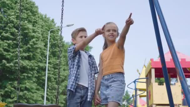 Cute boy and beautiful girl with long hair pointing away holding hands near the swing, smiling happily outdoors. A couple of happy children. Funny carefree kids in love. — Stock Video