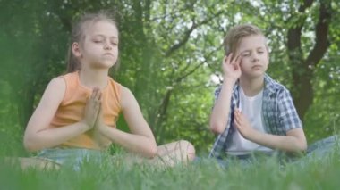 Portre oldukça sevimli küçük kız ve yakışıklı çocuk çim meditasyon oturuyor. Çocuklar yoga da var. Çocukların ruhsal gelişimi. Yaz tatili. Alt görünüm