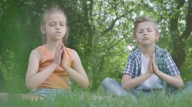 Güzel küçük bir kız ve çimenlerin üzerinde meditasyon yapmakta olan yakışıklı çocuk. Çocuklar yoga da var. Çocukların ruhsal gelişimi. Yaz tatili. Alt görünüm