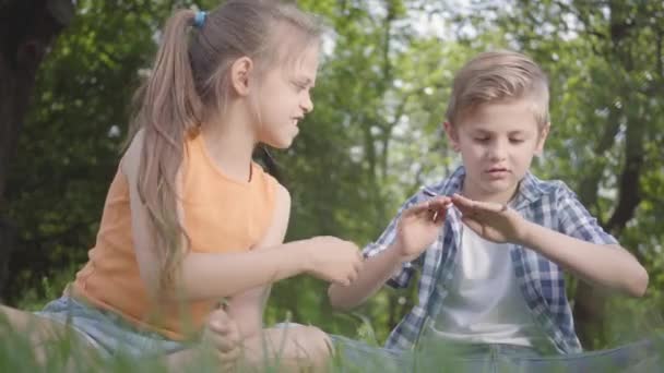 Két vicces gyerek ül a fűben a parkban játszik. A fiú játszik egy bug, lány megpróbálta elvenni. Egy pár boldog gyereknek. Vicces gondtalan gyerekek pihenni a szabadban. Alulnézet — Stock videók