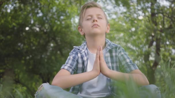 Bonito menino bonito sentado na grama meditando. Pratica ioga infantil. O desenvolvimento espiritual das crianças. Lazer de verão . — Vídeo de Stock