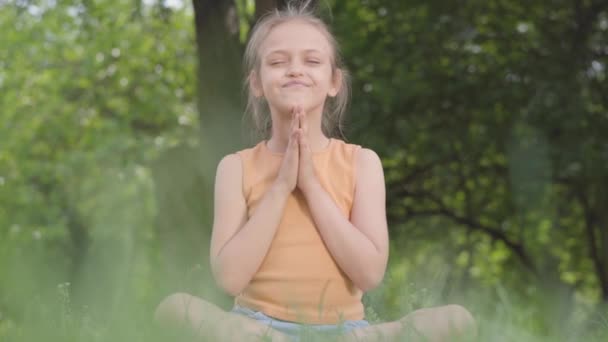 Söt liten ute flicka sitter på gräset mediterar. Barn praxis yoga. Sommar fritid. — Stockvideo