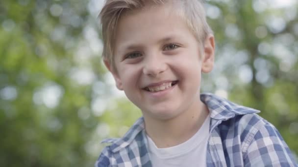 Zbliżenie portret cute przystojnego chłopca w kratkę patrząc na kamerę siedzi w parku. Summertime wypoczynek na świeżym powietrzu. Piękne dziecko studiując pod drzewami — Wideo stockowe