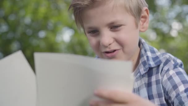 Close up portrait of cute handsome boy in checkered shirt looking at the sheets of paper in the park. Summertime leisure outdoors. Lovely kid studying under the trees — Stock Video