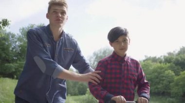 Sevimli genç baba parkta scooter binmek için oğlu öğretiyor. Adam çocuğu destekliyor, dengesini korumasına yardım ediyor. Baba ve bir çocuk dışarıda birlikte vakit geçirirler. Ön görünüm