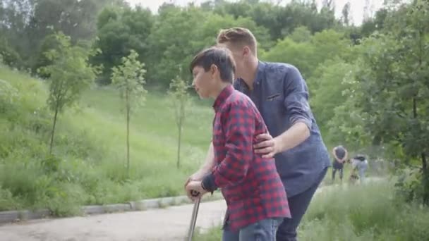 Vader leert zijn zoon om de scooter in het park te rijden. De man ondersteunt de jongen, hem helpen om evenwicht te houden. Vader en een kind tijd samen doorbrengen buiten — Stockvideo