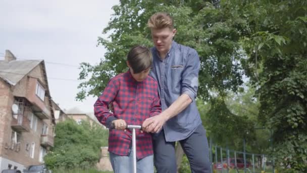 Portret schattige jonge vader leert zijn zoon om de scooter in het park te rijden. De man ondersteunt de jongen, hem helpen om evenwicht te houden. Vader en een kind brengen samen tijd door in de buitenlucht. Vooraanzicht — Stockvideo