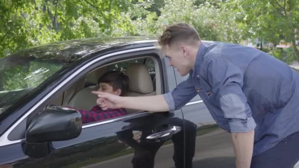 Junger Vater bringt seinem Sohn bei, das Auto zu fahren, das in der Nähe des Fahrzeugs steht. Der Junge sitzt auf dem Fahrersitz und hört Papa zu. Kind und Vater verbringen Zeit miteinander. Mann gibt Wissen an Sohn weiter — Stockvideo