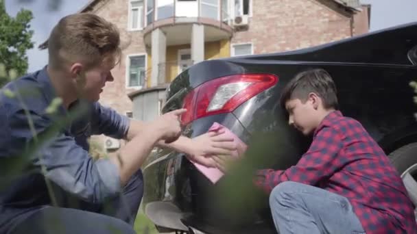 年轻的父亲教他的儿子关心汽车。 这个男人和一个男孩用抹布的特写擦拭机器。 一个男人把知识传给一个儿子，教他小心开车 — 图库视频影像
