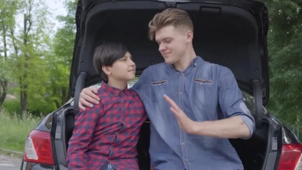 El joven padre y su hijo sentados en la parte trasera del coche al aire libre. El hombre abrazando al niño, la gente mirándose entre sí que a la cámara, sonriendo. Padre y un niño pasan tiempo juntos al aire libre — Vídeos de Stock