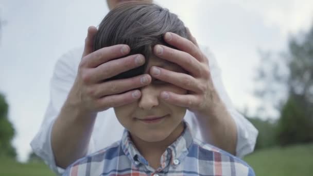 Retrato de cerca del chico guapo mirando a la cámara, su padre o hermano mayor cubriéndose los ojos por detrás. El niño sonriendo felizmente. Vacaciones familiares al aire libre. Padre y un niño pasan tiempo — Vídeo de stock