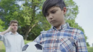Futbol toplarını ön planda tutan kameraya bakan üzgün bir çocuğun portresi. Genç meşgul baba arka planda cep telefonu ile konuşuyor.