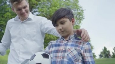 Parkta ön planda futbol topu tutan çocuk. Genç baba gelip çocuğa sarılıyor, kameraya bakıyor ve gülümsüyor. Aile eğlencesi açık havada. Baba ve çocuk birlikte vakit geçiriyorlar..