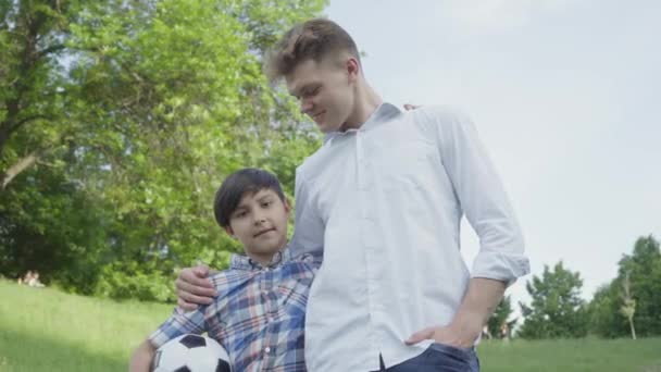 Adorable niño sosteniendo pelota de fútbol abrazándose con el padre mirando en cámara en el parque. Vacaciones familiares al aire libre. El chico jugando al fútbol con su padre. Padre y un niño pasan tiempo juntos. Vista inferior — Vídeos de Stock