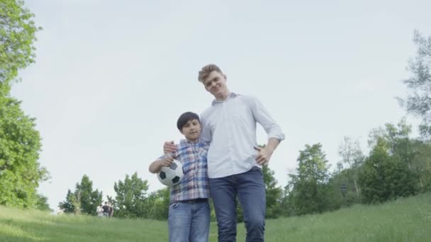 Il bambino che tiene il pallone da calcio che abbraccia con il padre che guarda in macchina fotografica nel parco. Tempo libero in famiglia all'aperto. Il ragazzo e suo padre mostrano i pollici in alto. Papà e un bambino passano del tempo insieme . — Video Stock