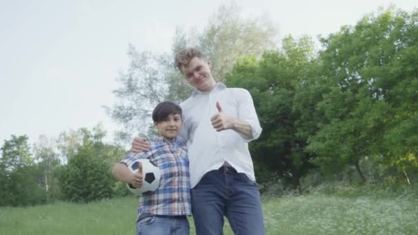 Portret jongen en zijn vader tonen duimen omhoog. Het kind het houden van Soccer Ball knuffelen met de vader op zoek in de camera in het Park. Familie vrije tijd buitenshuis. Vader en een kind brengen samen tijd door. — Stockvideo