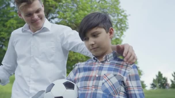 公園の前景でサッカーボールを持っている子供。若いお父さんが来て、少年を抱きしめ、カメラに向かって見て微笑んでいました。屋外で家族のレジャー。父と子供は一緒に時間を過ごす. — ストック動画