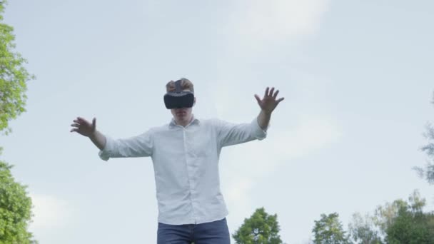 Jonge man in virtual reality headset spreidde zijn armen naar de zijkant, het nabootsen van de vlucht van een vliegtuig in het park genietend van het realistische beeld. Moderne technologieën. De man die een videospel speelt — Stockvideo