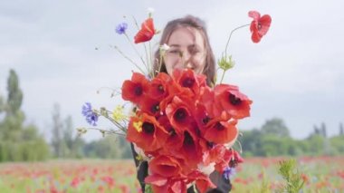 Bir haşhaş tarlasında güzel genç kız elinde çiçek buketi tutan kameraya bakıyor. Doğayla bağlantı. Yeşil ve kırmızı uyum. Doğada eğlence.