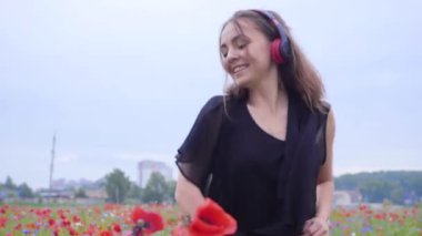 Müzik dinleyerek ve bir haşhaş alanında mutlu bir şekilde gülümseyen dans kulaklık takan güzel genç kadın portresi. Doğayla bağlantı. Doğada boş zaman. Çiçek açan haşhaşlar. Özgürlük.