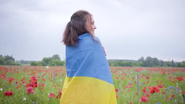 Hübsches Mädchen, das in einem Mohnfeld steht, das mit der Fahne der Ukraine bedeckt ist. Verbindung mit der Natur, Patriotismus. Freigeist, Emotionen, Leidenschaft. Freizeit in der Natur. Blühender Mohn. Freiheit. Zeitlupe. — Stockvideo
