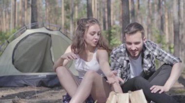 Portre genç adam ormanda bir yangın kindling sevimli genç kadın yakınında otururken. Sevgi dolu bir çift dışarıda dinleniyor. Kamp kavramı. Eğlence ve doğaya yolculuk.