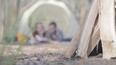 Portre genç adam güzel genç kadın ormanda çadırda birbirlerine yakın kitap okuma yatarken. Ön planda yakacak odun. Dışarıda eğlenen sevgi dolu bir çift. Kara zeminde bulanıklaştırma.