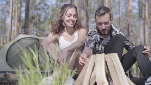 Porträtt stilig ung man Kindling en brand i skogen medan bedårande ung kvinna sitter nära. Kärleksfullt par som vilar utomhus. Begreppet Camping. Fritid och resa till naturen. — Stockvideo