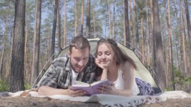 Portre genç adam ve güzel genç kadın ormanda çadırda kitap okuma birbirlerine yakın yatarken. Dışarıda eğlenen sevgi dolu bir çift. Kamp kavramı. Eğlence ve doğaya yolculuk.