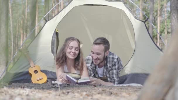 Retrato jovem mulher muito jovem deitado perto um do outro na tenda na floresta lendo o livro. Fogão em primeiro plano. Casal amoroso se divertindo ao ar livre. Conceito de acampar. Viagem . — Vídeo de Stock