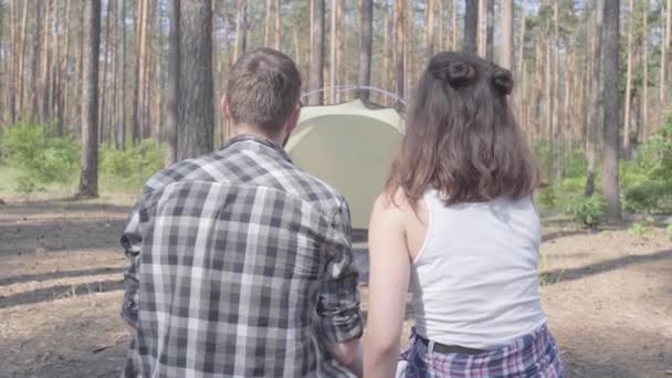 Retrato joven hombre y mujer sentados uno al lado del otro con la espalda a la cámara en el bosque. La tienda en el fondo. El hombre se gira y mira a la cámara. Una pareja de enamorados descansando al aire libre. Unidad con — Vídeos de Stock