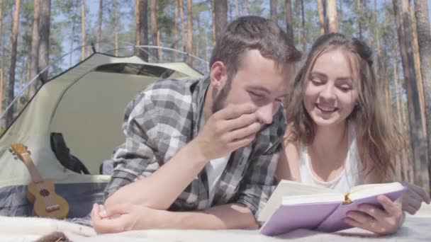 肖像画のあごひげの男とかわいい若い女性は、本を読んで森のテントの中で互いに近くに横たわっています。屋外で楽しいカップルを愛する。キャンプのコンセプト。レジャーと自然への旅. — ストック動画