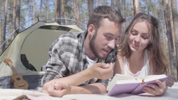 초상화 수염 남자와 사랑스러운 젊은 여자는 책을 읽고 숲에서 텐트에 서로 가까이 누워. 사랑스러운 부부는 야외에서 즐거운 시간을 보내고 있습니다. 캠핑의 개념. 자연으로의 레저 및 여행. — 비디오