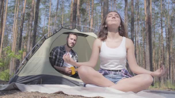 若い女性が前景で瞑想しながら、テントの中でウクレレを弾いている男。愛するカップルは、屋外で休んでいます。キャンプのコンセプト。レジャーと自然への旅. — ストック動画