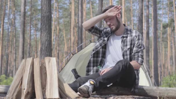 Young handsome man sitting in front of pieces of wood for the fire, drinking water from the plastic bottle. The tent on the background. Unity with wild nature. The guy is camping — Stock Video