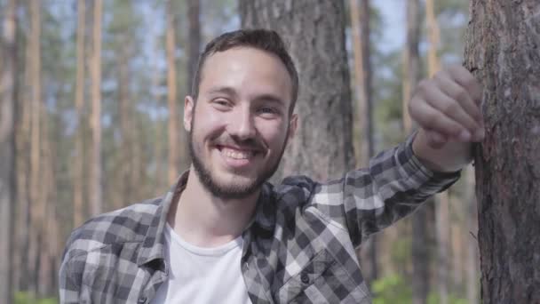 Retrato de un joven guapo en el bosque de pinos, mirando a la cámara y sonriendo de cerca. Unidad con la naturaleza salvaje. El tipo descansando al aire libre — Vídeos de Stock