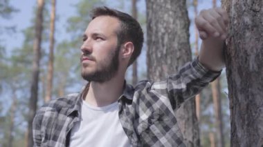 Çam ormanında yakışıklı sakallı genç adamın portresi, kameraya bakıyor ve yakın çekimgülümsüyor. Vahşi doğayla birlik. Dışarıda dinlenen adam.