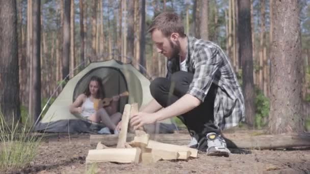 Knappe man in een geruite overhemd bereidt brandhout om een brand buitenshuis te maken. Het meisje zit in een tent en speelt de ukulele of gitaar. Concept van kamperen. Vrije tijd en reis naar de natuur. — Stockvideo