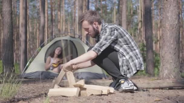 Młody mężczyzna rozpala ogień na pierwszym planie w lesie, podczas gdy całkiem młoda kobieta gra na ukulele na tle w namiocie. Kochająca para odpoczynku na zewnątrz. Jedność z naturą — Wideo stockowe